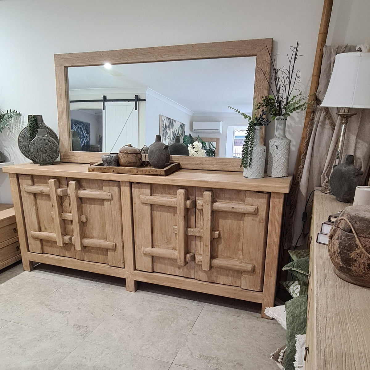 A light natural wood sideboard cabinet with a large matching wood-framed mirror, styled in a home interior with decorative vases and plants.