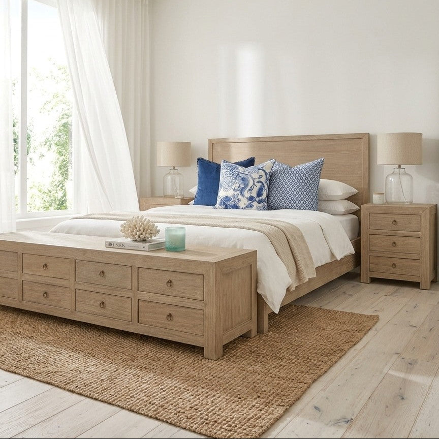 A bright, modern bedroom featuring a light natural wood bed frame, matching nightstands, and a wooden bench at the foot of the bed. The room is styled with white bedding, blue patterned accent pillows, a natural jute rug, and soft natural light coming from a window with sheer white curtains.